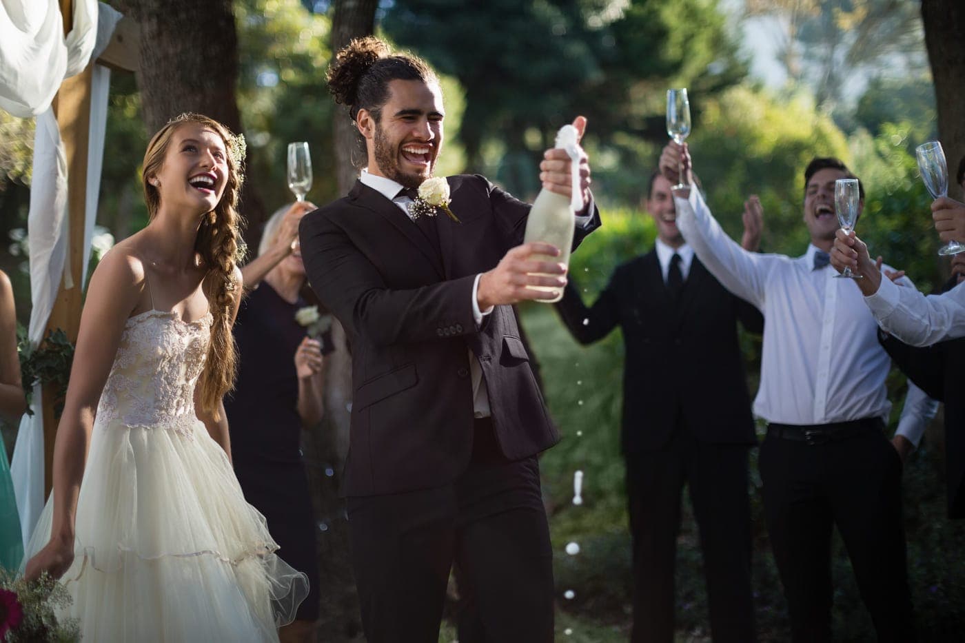 Bräutigam öffnet schwungvoll eine Sektflasche, während die Braut und feiernde Hochzeitsgäste im Hintergrund jubeln, ein Highlight der Hochzeit.