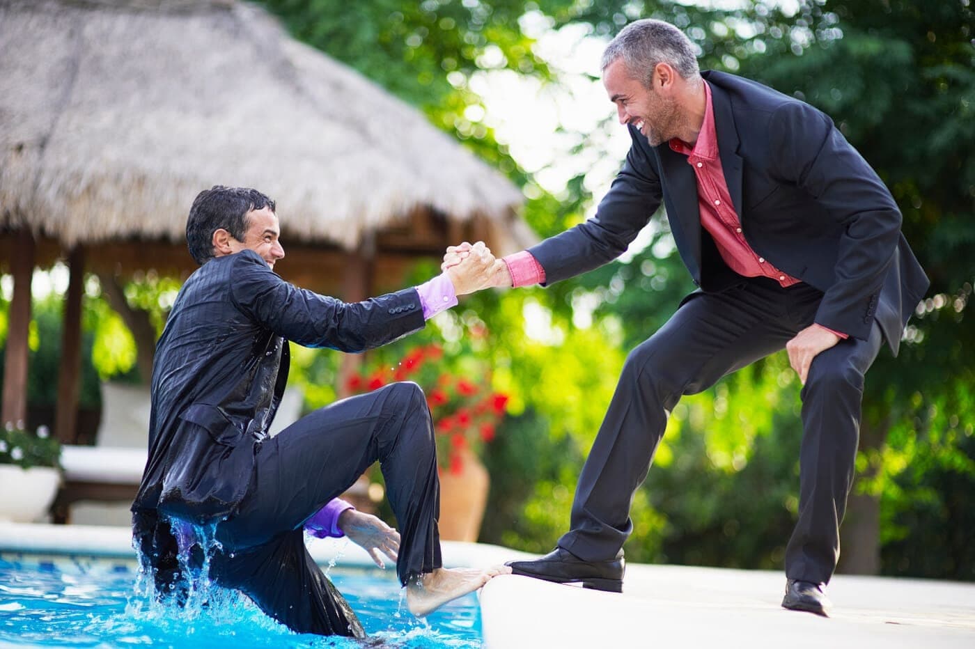Lachender Gast im Anzug wird von einem anderen Gast aus dem Pool gezogen, ein Beispiel für ausgelassene Stimmung und lustige Ideen auf einer Hochzeit.