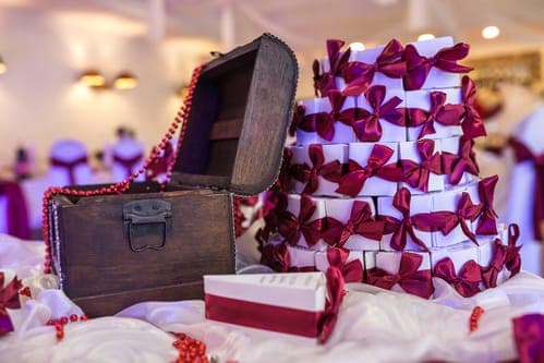 Pyramide aus weissen Gastgeschenken zur Hochzeit mit violetten Schleifen, gestapelt neben einer offenen Holztruhe als Give-away für die Gäste.