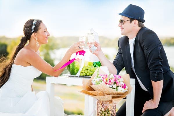 Entspanntes Brautpaar stösst an einem weissen Tisch mit Sektgläsern an, ein Moment zum Durchatmen im Hochzeitsprogramm nach der Zeremonie.