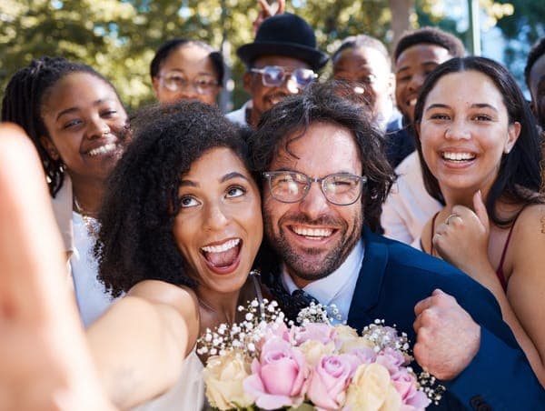 Brautpaar macht ein fröhliches Selfie mit lachenden Hochzeitsgästen im Hintergrund im Freien.