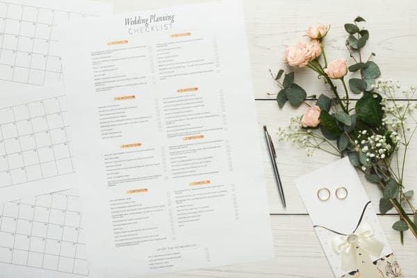 A printed wedding planning checklist and calendar pages beside a pen, pale pink roses and two gold rings on a white wooden table