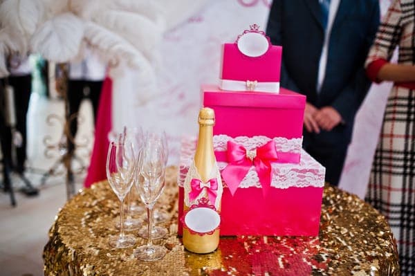 Pinker Geschenkbox-Turm mit Spitzen-Deko und goldene Sektflasche auf dem Gabentisch.