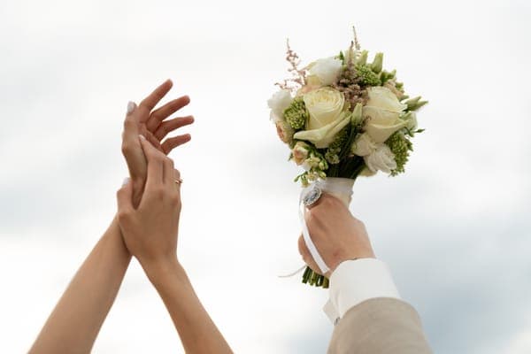 Eine Hand hält einen weissen Brautstrauss hoch, andere Hände strecken sich danach vor hellem Himmel. Symbolischer Augenblick für Hochzeitswünsche und Feier, welche sich erfüllen sollen.