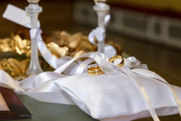 Zwei goldene Eheringe, die dekorativ auf einem weissen Satin Ringkissen mit Schleifen liegen.