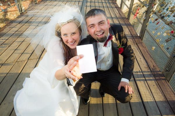 Braut und Bräutigam knien auf einem Holzsteg und halten eine leere Hochzeitskarte in die Kamera; Platz für persönliche Hochzeitswünsche und einen Hochzeitswunsch.