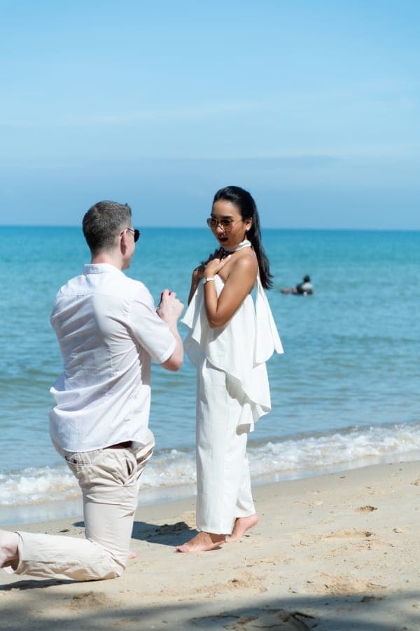 A man kneeling in the sand in front of his surprised partner, who is dressed in white, with the blue sea behind them