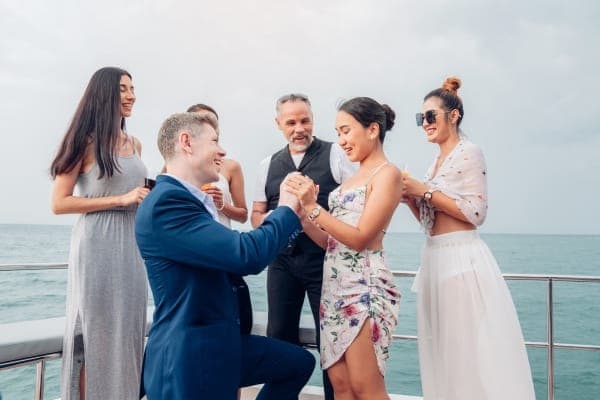 A man in a blue suit on one knee in front of his partner on a boat, with friends watching in the background