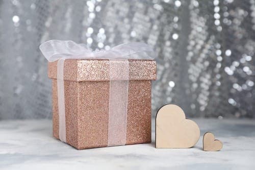 Roségoldene Glitzer Geschenkbox mit weisser Schleife und zwei Holzherzen vor Bokeh Hintergrund