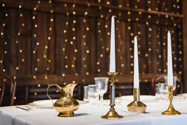 Weisse Stabkerzen in Messinghaltern auf einem Hochzeitstisch vor einer rustikalen Holzwand mit unscharfer Lichterkette.