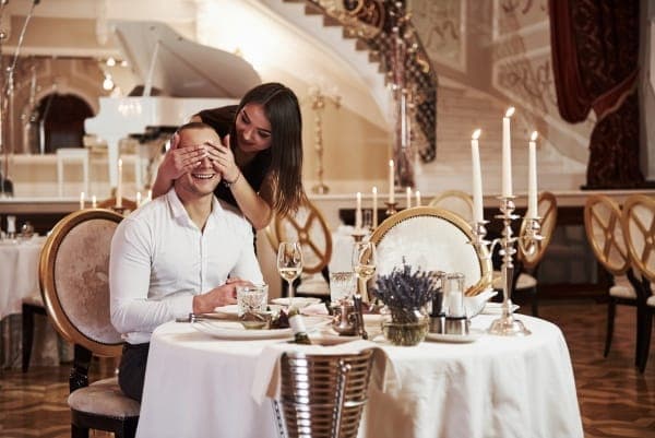 Überraschung beim romantischen Dinner: Eine Frau hält ihrem Partner in einem eleganten Restaurant bei Kerzenschein die Augen zu, passend als Idee für einen intimen Heiratsantrag.