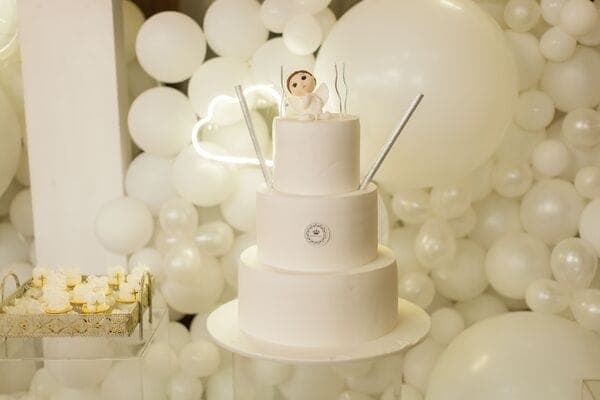 Dreistöckige weisse Torte mit Engel-Topper vor weisser Ballonwand.