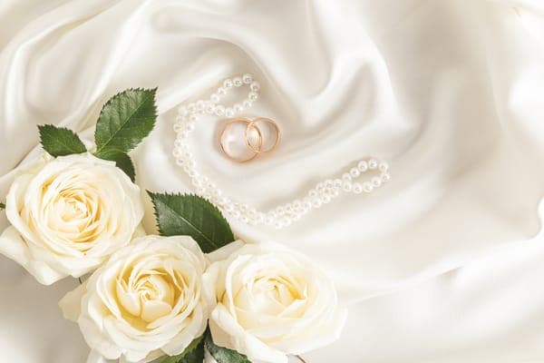 Goldene Eheringe in Perlen-Herzform neben weissen Rosen auf Satin.