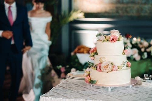 Dreistöckige Hochzeitstorte mit rosa Blumen auf gedecktem Tisch, im Hintergrund das Brautpaar bei der Feier.