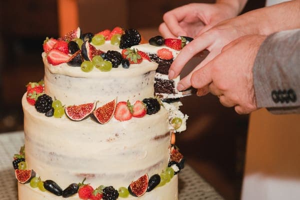 Zwei Hände schneiden ein Stück aus einer Semi-Naked-Hochzeitstorte mit Beeren, Feigen und Trauben.