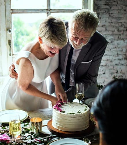Ein älteres Brautpaar schneidet lachend eine Hochzeitstorte an einem festlich gedeckten Tisch vor einem grossen Fenster an.