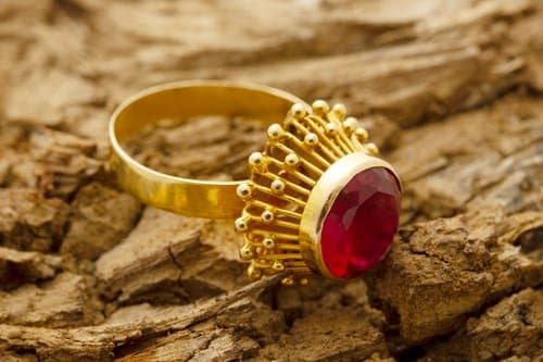 Goldener Ring mit grossem roten Edelstein liegt auf rauer Baumrinde; der Fokus liegt auf dem funkelnden Stein.