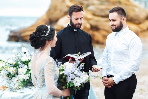 Eine Trauung am Meer: Ein Geistlicher in schwarzer Robe liest aus einem Buch, gegenüber steht ein Bräutigam im weissen Hemd. Die Braut in hellblauem Kleid hält einen grossen weissen Blumenstrauss, im Hintergrund sind Felsen und Wellen.