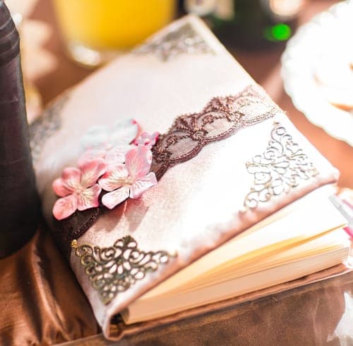 Ein verziertes Gästebuch mit glänzendem Stoffeinband, Spitze, rosa Blumen und Metallecken auf einem Tisch.