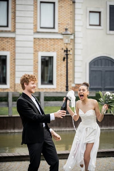 Ein Mann im schwarzen Anzug spritzt Sekt aus einer Flasche; eine jubelnde Braut im kurzen weissen Kleid hält einen Blumenstrauss. Im Hintergrund stehen Häuser am Wasser.