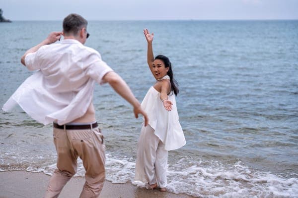 Am Strand öffnet ein Paar die Arme zueinander: eine Frau in weissem Outfit steht barfuß am Wasser, ein Mann in hellem Hemd dreht sich zu ihr. Hinter ihnen liegt das ruhige Meer bis zum Horizont.