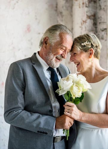 Ein älteres Brautpaar lehnt die Stirnen aneinander und lacht, während die Braut einen Strauss weisser Rosen in den Händen hält.