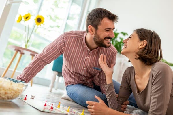 Paar sitzt am Boden und spielt Brettspiel; sie lachen miteinander, Spielsteine und eine Schüssel Popcorn liegen daneben.