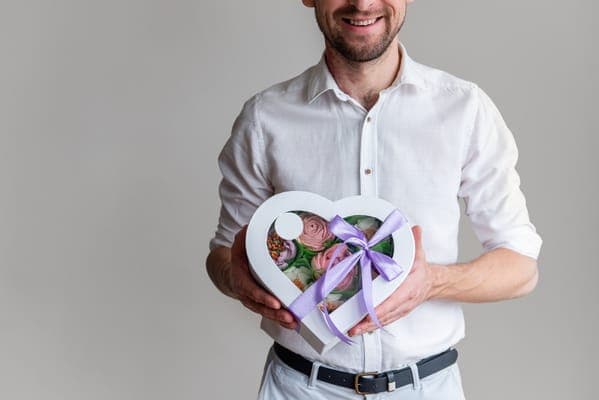 Mann im weissen Hemd hält eine herzförmige Geschenkbox mit lila Schleife