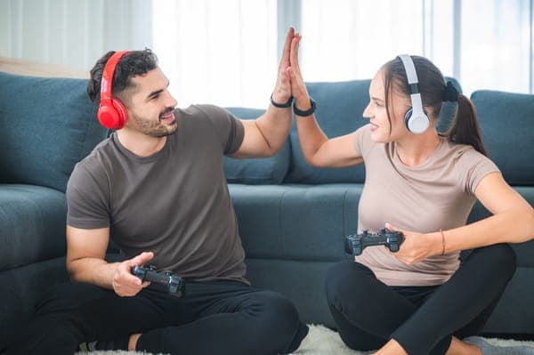 Paar mit Kopfhörern gibt High Five beim Gaming; beide sitzen vor dem Sofa und halten Controller in der Hand.