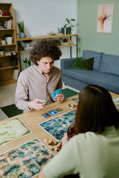 Mann und Frau spielen Brettspiel am Tisch; Spielpläne, Karten und Spielfiguren liegen aus, beide wirken konzentriert.
