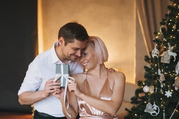 Paar neben einem geschmückten Weihnachtsbaum hält ein kleines Geschenk mit Schleife.
