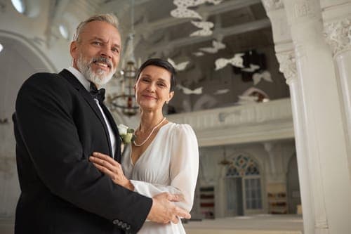 Älteres Hochzeitspaar umarmt sich in einer hellen Kirche. Er trägt Smoking, sie ein schlichtes weisses Kleid mit Perlenkette