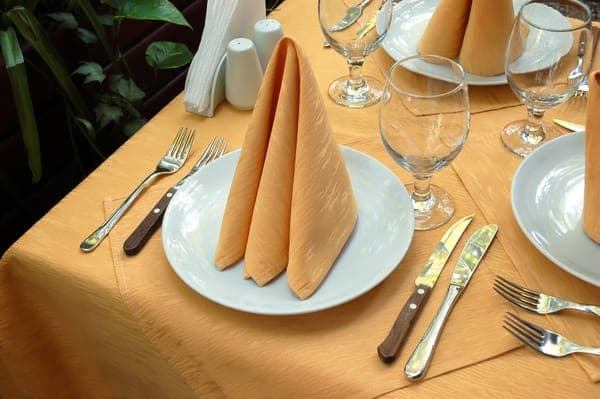 Orange Serviette zur Zeltform gefaltet auf Teller, umgeben von Gläsern und Besteck auf gedecktem Tisch.