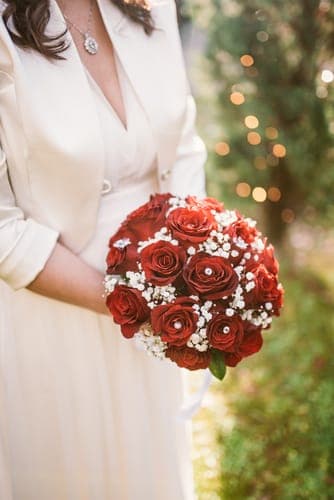 Person im weissen Outfit hält einen Strauss aus roten Rosen und Schleierkraut vor unscharfem Grün mit Lichtern.