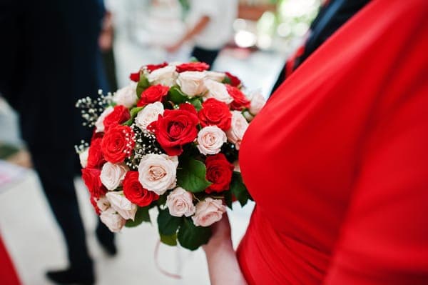 Hochzeitsgast im roten Kleid hält einen runden Brautstrauss aus roten und zartrosa Rosen mit Grün.