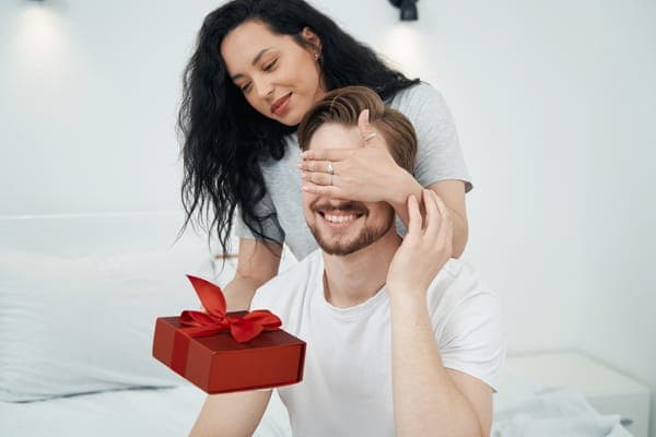 Frau überrascht lachenden Mann, hält ihm die Augen zu und reicht eine rote Geschenkbox