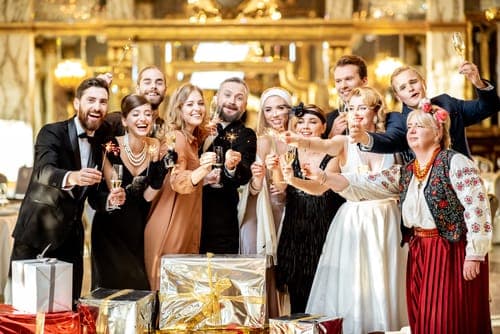 Gruppe festlich gekleideter Hochzeitsgäste stösst mit Sektgläsern an und hält Wunderkerzen; vorne liegen mehrere eingepackte Geschenke und im Hintergrund ein goldener Festsaal.