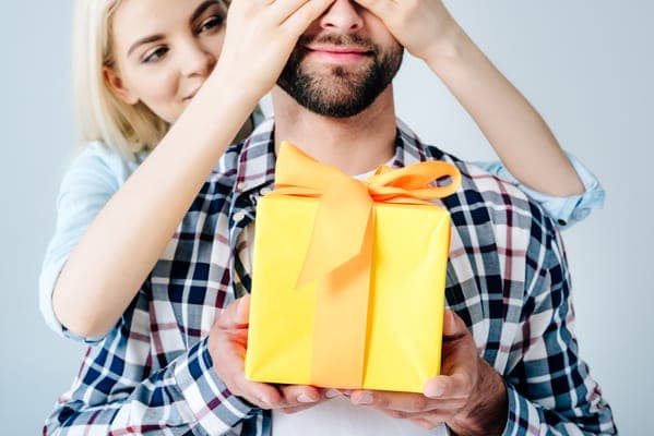 Frau hält einem Mann die Augen zu und überreicht ein gelbes Geschenk mit orangefarbener Schleife