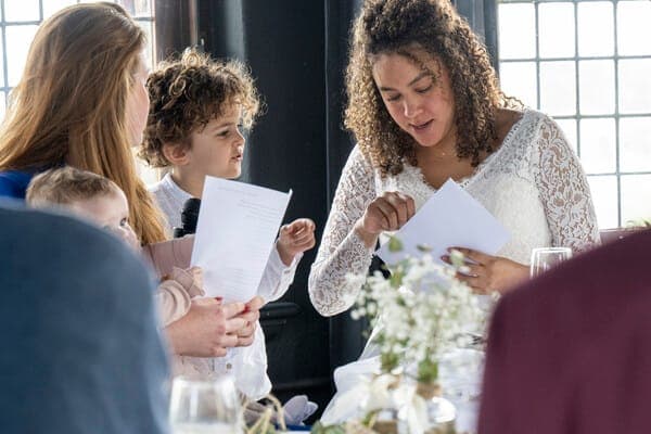 Braut im Spitzenkleid öffnet einen Umschlag am Tisch, daneben eine Frau und zwei Kinder mit Papieren.