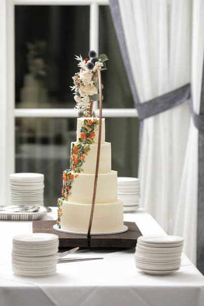 Vierstöckige Hochzeitstorte auf Desserttisch, mit bunten Blüten-Dekoren und floralem Bogen; Tellerstapel daneben.