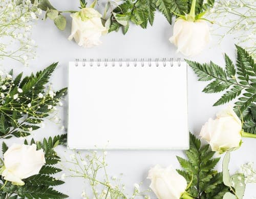 Leeres spiralförmiges Notizbuch auf hellem Hintergrund, dekorativ eingerahmt von weissen Rosen, Farn und feinen weissen Blüten.