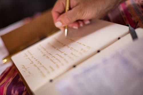 Eine Hand, die mit einem goldenen Stift in ein aufgeschlagenes Gästebuch schreibt; der Text ist unscharf.