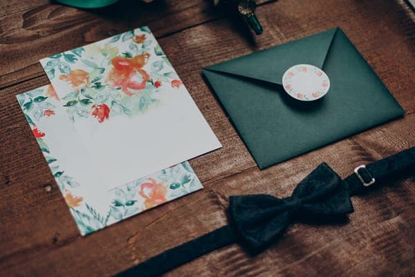 Hochzeitseinladung-Set auf Holztisch: Aquarellkarte mit Blumenrand, dunkelgrüner Umschlag mit rundem Siegel sowie schwarze Fliege.
