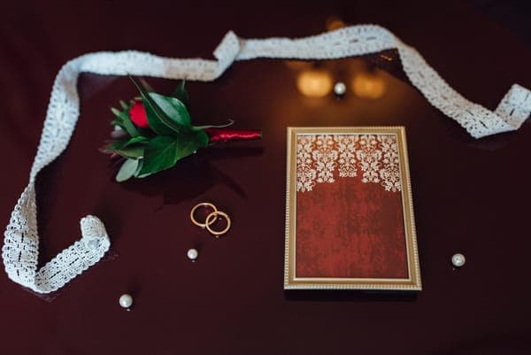 Bordeauxfarbene Hochzeits-Deko in Detailaufnahme: Spitzenband als Rahmen, roter Rosen-Anstecker, zwei Eheringe und Perlen neben einer rot-goldenen Karte/Box auf dunklem Tisch.