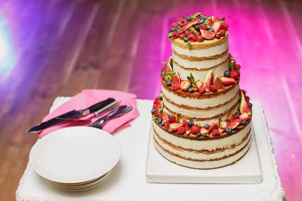 Dreistöckige Naked-Hochzeitstorte mit Beeren auf Tisch; daneben Tellerstapel und Besteck auf pinken Servietten.