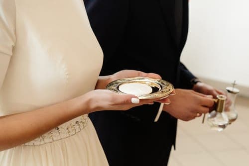 Braut im weissen Kleid hält eine Kerzenschale; daneben hält der Bräutigam in dunklem Anzug ein kleines Glas