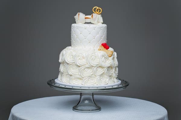 Zweistöckige weisse Hochzeitstorte mit Rautenprägung und Rosen-Creme, auf Glasplatte vor grauem Hintergrund.