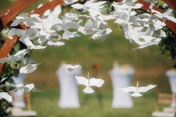 Viele weisse Papier-Vögel hängen als Hochzeitsdeko unter einem Holzbogen; im Hintergrund stehen Stehtische auf einer Wiese.