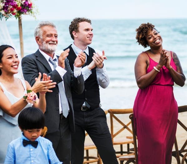 Hochzeitsgäste applaudieren bei einer Strandhochzeit, Meer und Wellen im Hintergrund, festliche Stimmung.