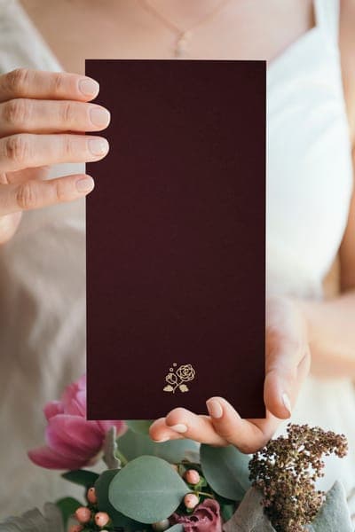 Hände halten bordeauxfarbene Einladungskarte: goldenes Rosenmotiv unten; im Vordergrund ein Blumenstrauss mit Eukalyptus und rosa Blüten, eleganter Hochzeitspapeterie-Look.